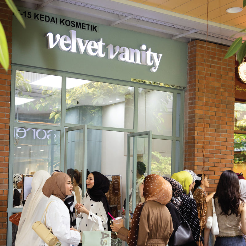 A group of women wearing modest clothing and headscarves stand outside the velvet vanity cosmetics store, with the storefront, glass doors, and brick wall visible in the background.